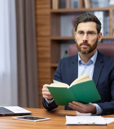 Portrait of a young businessman working in the office at a laptop and holding a book. He looks seriously and confidently into the camera.