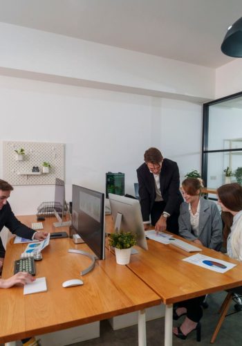 A group of five professionals engages in a business meeting in a contemporary office. They are discussing project plans while reviewing documents and using computers.