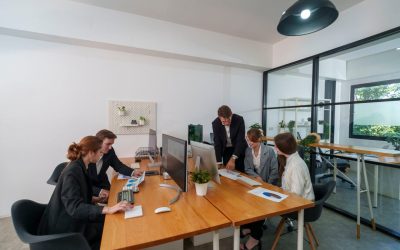 A group of five professionals engages in a business meeting in a contemporary office. They are discussing project plans while reviewing documents and using computers.