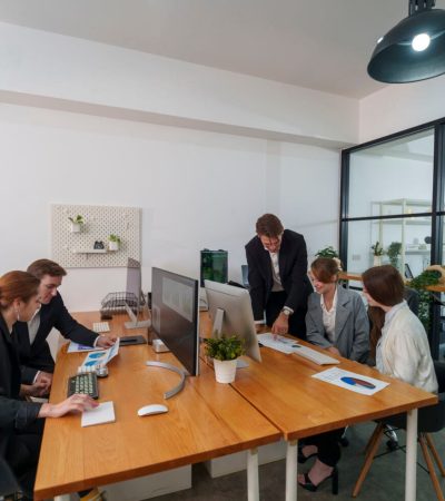 A group of five professionals engages in a business meeting in a contemporary office. They are discussing project plans while reviewing documents and using computers.