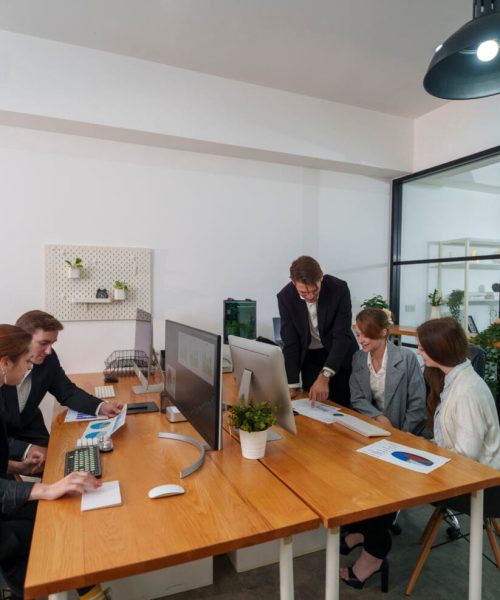A group of five professionals engages in a business meeting in a contemporary office. They are discussing project plans while reviewing documents and using computers.