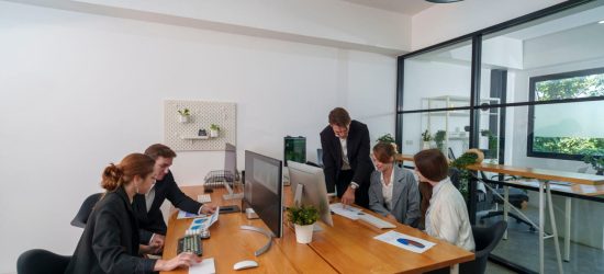 A group of five professionals engages in a business meeting in a contemporary office. They are discussing project plans while reviewing documents and using computers.